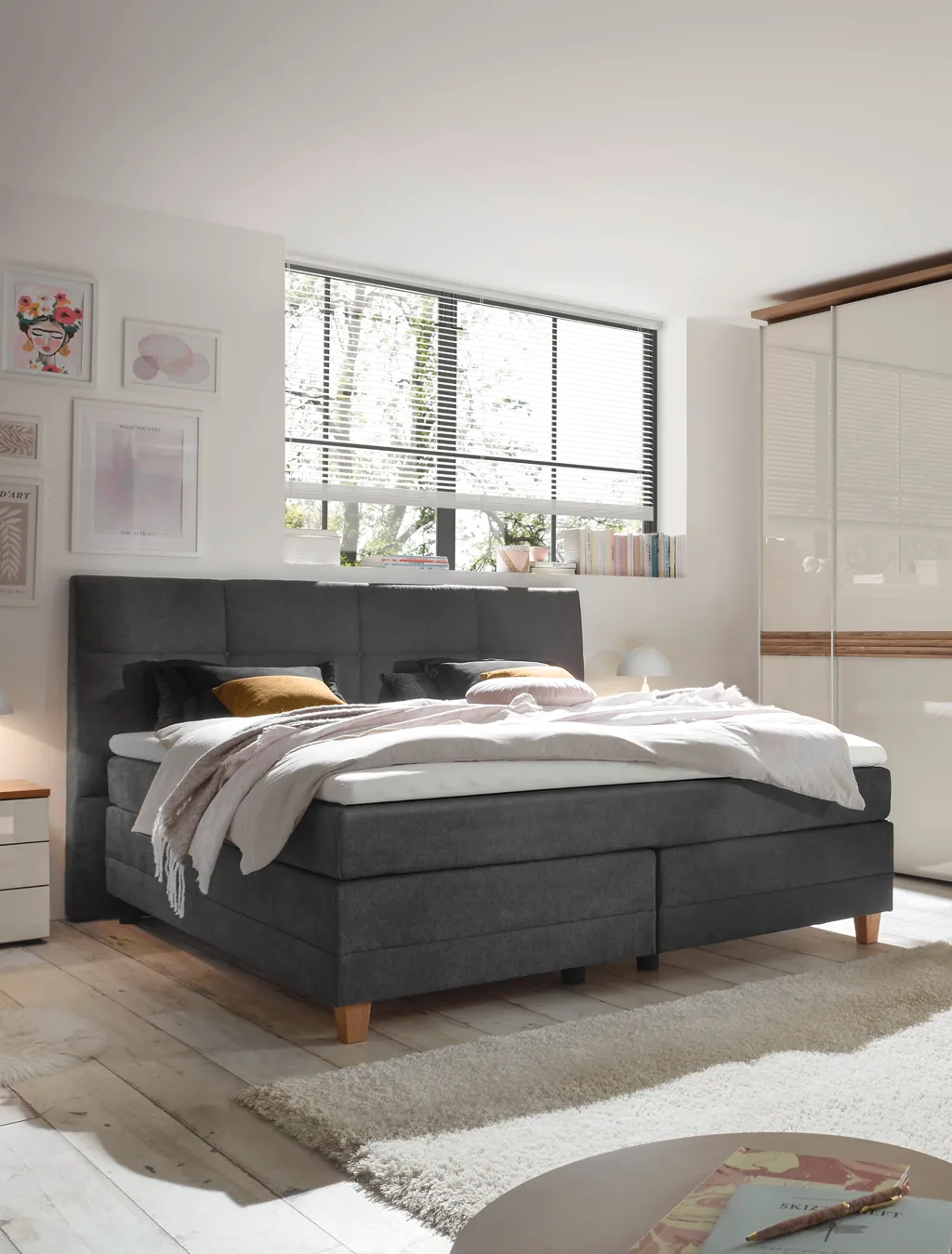 Modernes Boxspringbett in elegantem Grau mit Holzfüßen, Kopfteilpolsterung und heller Bettwäsche. Helles Schlafzimmer mit Deko-Bildern, Teppich und Kleiderschrank – ideal für Komfort und gesunden Schlaf.